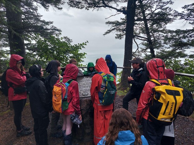 Camp kids in rain gear huddled around a commemorative plaque emplaced upon a stone block at Fort McGilvray in Caines Head State Recreation Area